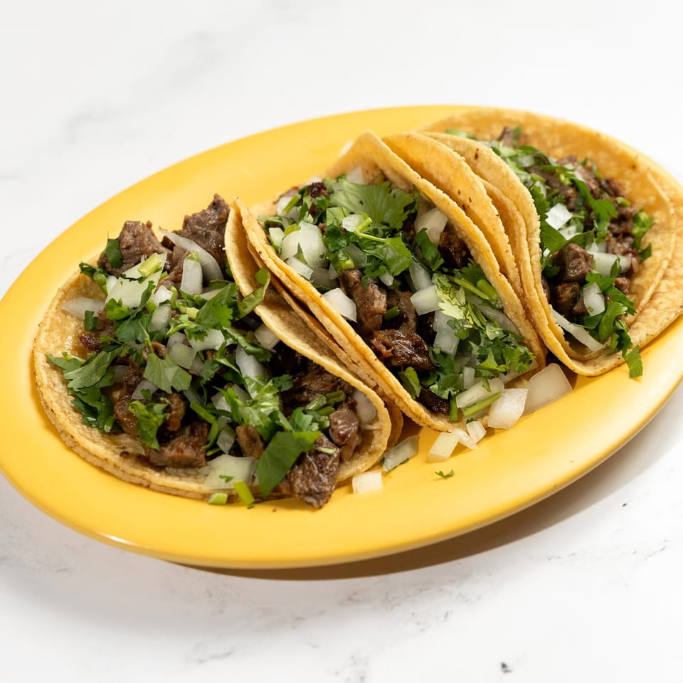 Three De Pasada Mexican street tacos with carne asada, cilantro, and diced onions on a yellow plate.