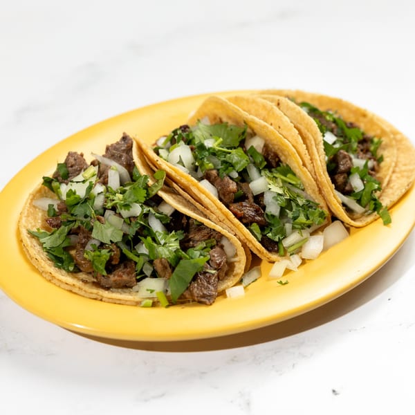 Three De Pasada Mexican street tacos with carne asada, diced onions, and cilantro on a yellow plate.